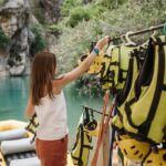 A Woman Getting a Helmet from a Rack, also yellow life jackets and a river is in the background for river rafting