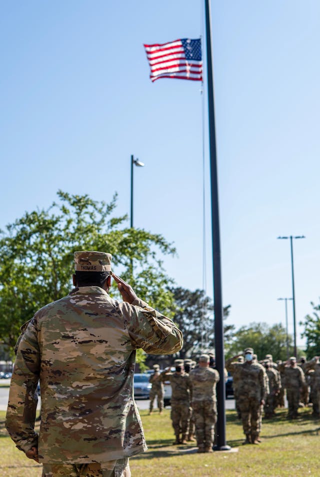 A group of soldiers in uniform salute the American flag during a daytime ceremony.